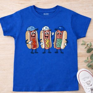 May include: Royal blue t-shirt with a cartoon illustration of three hot dogs wearing baseball caps with "KC" on them. Each hot dog has a different topping: ketchup, mustard, and relish. The shirt is made of a soft material.
