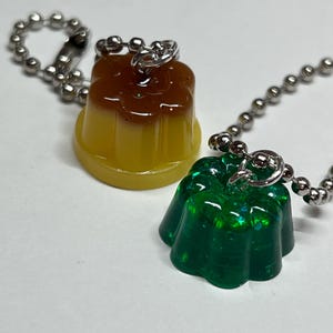 May include: Two resin charms shaped like desserts. One is a two-toned yellow and brown pudding, and the other is a green, glittery jelly. Both charms have silver ball chain keychains attached.