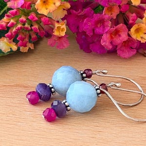 May include: A pair of dangle earrings featuring large, faceted, light blue round beads, smaller faceted purple beads, and tiny faceted magenta beads. The beads are accented with small silver-toned metal spacers and hang from curved silver-toned ear wires.