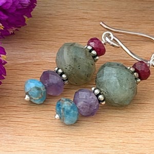 May include: A pair of dangle earrings featuring faceted beads in shades of gray, purple, blue, and red. Each earring is composed of a large gray stone, a smaller purple stone, and a small blue stone, accented with silver beads and silver hooks.