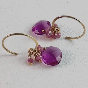 May include: A pair of gold hoop earrings featuring a large, faceted, teardrop-shaped purple gemstone and a cluster of smaller, round, pink gemstone beads suspended from the bottom of each curved gold wire hoop.