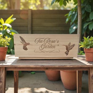 May include: A wooden planter box with the text "Tia Rosa's Garden" and hummingbird designs. The rectangular box is light brown and sits on a wooden table. Two small potted plants flank the box.