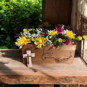 May include: A wooden planter box filled with yellow, purple, and pink flowers. A small wooden cross with the word "LOVE" is in front of the planter. The planter box has the words "Lupe's Garden" on the side.