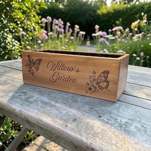 May include: A rectangular wooden planter box with a natural light brown finish. The front panel features a laser-engraved design of two butterflies and floral accents, with the text Willow's Garden centered between the butterfly illustrations.