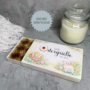 May include: A box of chocolates with a decorative lid featuring Easter-themed illustrations. The lid displays a lamb, basket of eggs, and text that reads "süße Ostergrüße für dich." A glass jar candle is in the background.