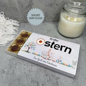 Könnte beinhalten: Eine weiße Schachtel Pralinen mit dem deutschen Text "Frohe Ostern" und einer Aquarellillustration eines Hasen, Ostereiern und einem Baum. Die Schachtel enthält mehrere runde Pralinen mit einer karamellfarbenen Füllung. Ein Glas mit einer weißen Kerze befindet sich im Hintergrund.