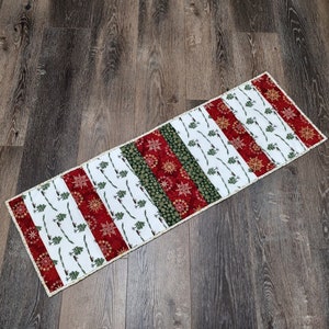 May include: A white table runner with alternating stripes of red, green, and white featuring a snowflake and tree design.