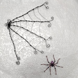 May include: A black wire spider web with decorative swirls and a beaded spider. The spider has a purple body and beaded legs. The web is made of thin, black wire and is a Halloween decoration.