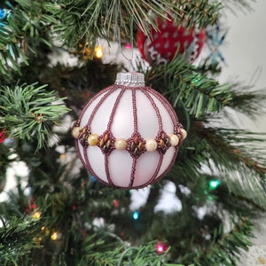 May include: A white Christmas ornament with a beaded design. The ornament is decorated with brown and gold beads, creating a geometric pattern.