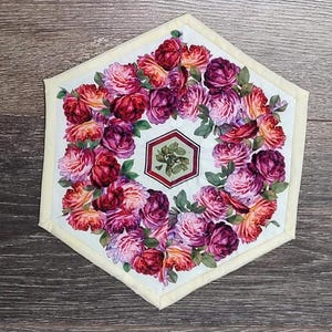 Könnte beinhalten: Ein sechseckiger, gesteppter Tischläufer mit weißem Hintergrund und einem Blumendesign mit rosa, roten und orangefarbenen Rosen. Die Mitte des Läufers zeigt ein kleineres sechseckiges Design mit grünen Blättern.