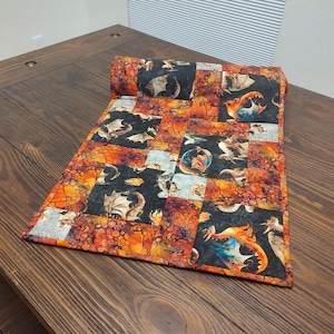 May include: A quilted table runner featuring a patchwork design with dragon motifs. The runner showcases a mix of black, orange, and gray squares, with dragon illustrations in various colors. The edges are neatly finished, and the runner is rolled up at one end.
