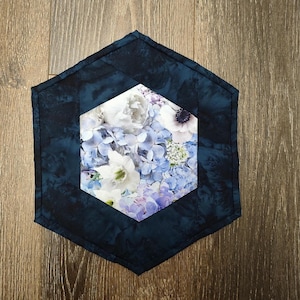 May include: A quilted, hexagonal placemat featuring a central floral design of white, blue, and purple flowers. The outer border is a dark blue fabric with a marbled effect. The placemat is placed on a wooden surface.