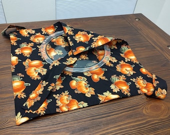 May include: A black fabric tote bag with a pumpkin pattern, featuring orange pumpkins and autumn leaves. The bag has a clear plastic ring handle and is displayed on a wooden surface.