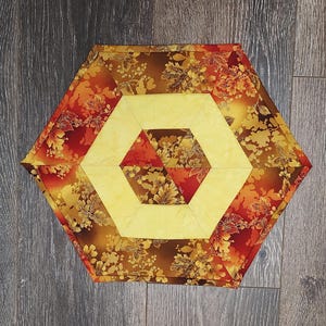 May include: A hexagonal table runner with a geometric pattern in shades of yellow, orange, and brown. The runner features a floral design with leaves and flowers.
