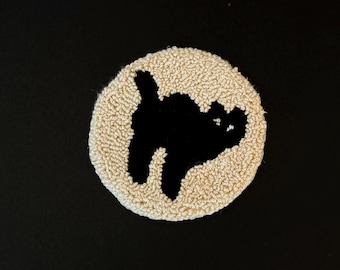 May include: A round, beige, hand-tufted coaster with a black cat silhouette in the center. The coaster has a textured surface and is set against a black background. The cat is depicted with its tail up.
