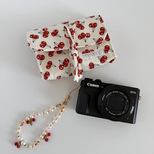 May include: A black Canon G7X camera with a cream-coloured case patterned with red cherries, and a beaded cherry-themed wrist strap. The case has the text "LOVE CI". The wrist strap features pearls and gold beads.
