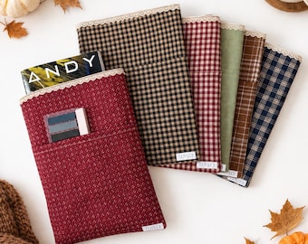 Pode incluir: Uma coleção de capas retangulares de tecido para livros com vários padrões, incluindo xadrez vermelho, preto e bege, verde sólido e tartan. Cada capa tem um acabamento em renda na borda superior e uma pequena etiqueta branca com a inscrição LOVECL.