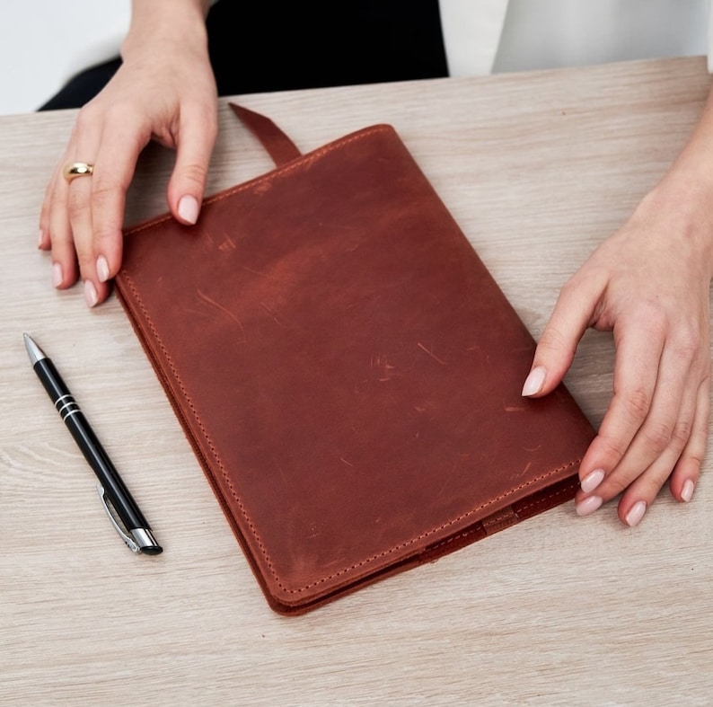 May include: A brown leather bound journal with a simple design. The journal is closed and lying flat on a wooden surface.