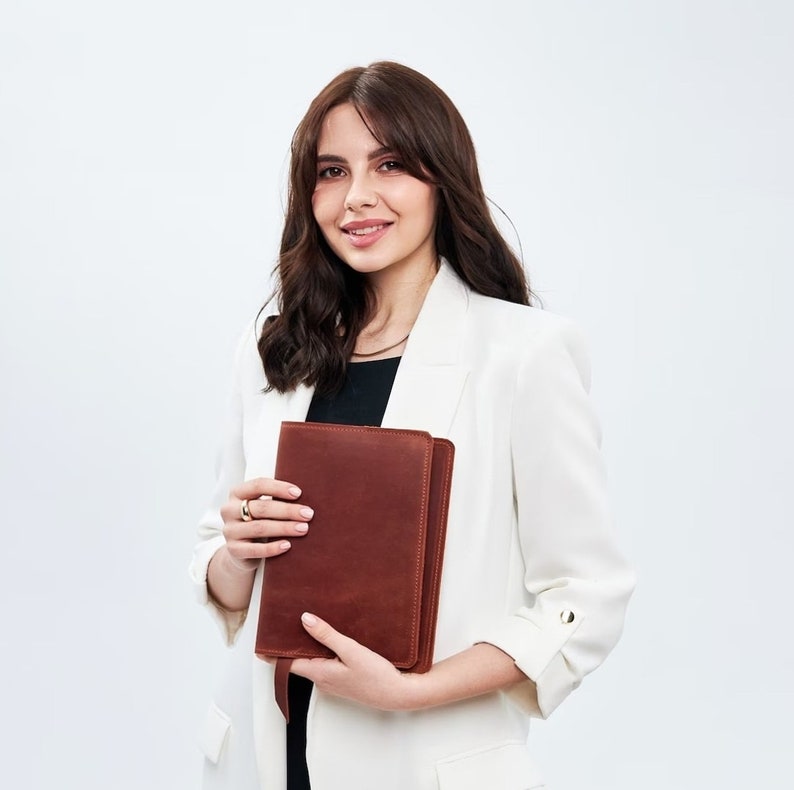 May include: A woman in a white blazer holds a brown leather bound journal. She is smiling at the camera.