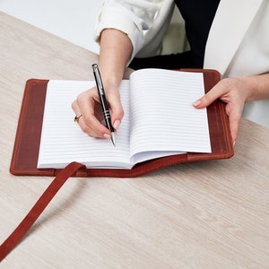 May include: A brown leather bound journal with a pen lying open on a white lined page. The journal has a brown leather strap and a red ribbon bookmark.