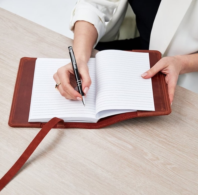 May include: A brown leather bound journal with a pen lying open on a white lined page. The journal has a brown leather strap and a red ribbon bookmark.