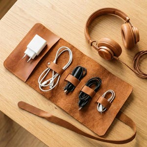 May include: A brown leather cable organizer with a white charger, white and black cables, and a brown leather strap. A pair of brown headphones rests on the wooden surface.