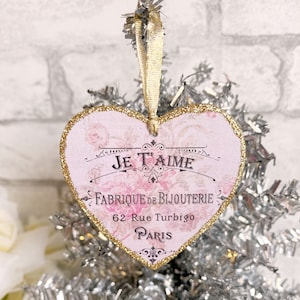Op de afbeelding: Roze hartvormige ornament met gouden glitterrand. Het heeft de tekst "Je t'aime" en "Fabrique de Bijouterie 62 Rue Turbigo Paris".