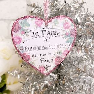 Op de afbeelding: Roze hartvormige ornament met een bloemenpatroon en de tekst "Je T'aime Fabrique de Bijouterie 62 Rue Turbigo Paris".