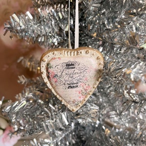 May include: A white heart-shaped Christmas bauble with gold trim and a floral design. The bauble has the text "Noël à Granit" in French, "Anglaise" in English, and "Paris" in French.