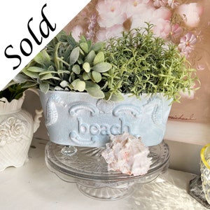 May include: A light blue ceramic planter with the word "beach" embossed on the front. The planter is filled with green plants and sits on a clear glass cake stand with a seashell on top.