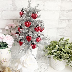 May include: A small silver artificial Christmas tree decorated with red heart-shaped ornaments. The tree is on a glass cake stand with a white rose on top.