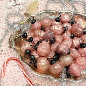 May include: A collection of pink glass Christmas baubles with black caps and silver metal hangers, displayed in a silver tray. The baubles have various shapes and textures. A red and white striped candy cane is in the foreground.