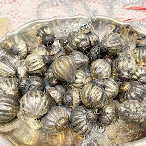 May include: A collection of vintage silver Christmas ornaments with ribbed and textured surfaces. The ornaments are arranged in a silver serving dish, creating a festive display. Each ornament has a small metal cap and loop for hanging.