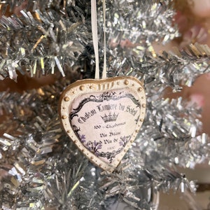May include: Heart-shaped Christmas tree decoration with a vintage wine label design. The ornament features the text "Chateau Lumiere du Soleil" and other details in black and purple. It has a cream-coloured border with gold accents and a white ribbon for hanging.