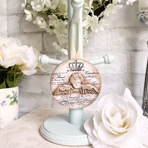 May include: A round, cream-colored ornament with a raised image of an angel with wings. The ornament has a gold crown and the text "Assigned for the Piano Soiree" in a decorative script.