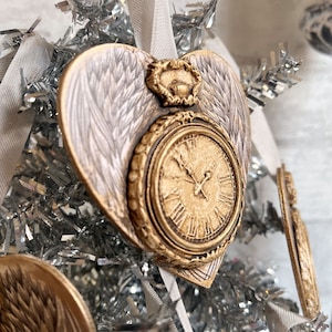May include: A gold heart-shaped ornament with a clock face and angel wings. The ornament has a white background and gold accents.