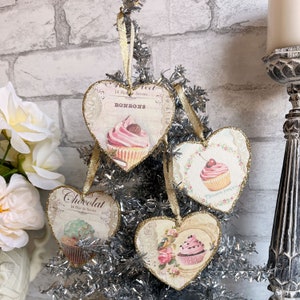 May include: Three pink and white heart-shaped decorations with gold ribbon and a silver and white Christmas tree in the background. Each decoration features a different cupcake design with text that reads "Bonbons", "Chocolat", and "Patisserie Francaise".