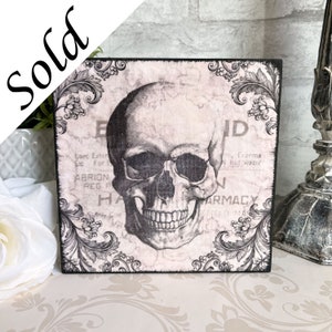 May include: A square wooden sign with a black and white skull graphic and ornate floral border. The sign has a distressed, vintage look and features the text "Esec Exter Etc For ABRION REG P HA N ID m, Eczema, hot water ARMACY".