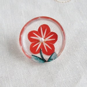 Op de afbeelding: Een doorzichtige glazen knoop met een rood en wit bloemenontwerp. De bloem heeft een wit centrum en rode bloemblaadjes met witte strepen. De knoop heeft een groene steel en bladeren.