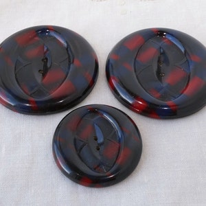 Puede incluir: Tres botones de plástico en un patrón de remolino rojo y azul. Los botones son redondos y tienen dos agujeros.