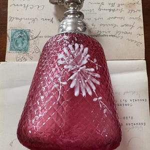May include: A vintage cranberry-coloured perfume bottle with a silver-toned atomizer. The bottle has a diamond pattern and a white floral design. The bottle is on top of old letters and postcards.
