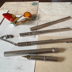 May include: A collection of antique writing instruments, featuring several silver-coloured pens and a fountain pen with its nib visible. The items are arranged on a selection of aged postcards with handwritten text and stamps. A decorative mushroom-shaped brooch is also present.
