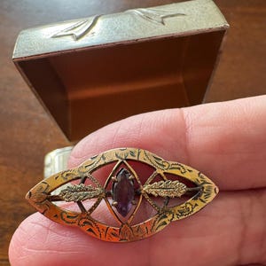 Op de afbeelding: Een vintage goudkleurige broche met een ovale vorm, met een amethistkleurige edelsteen in het midden. De broche heeft ingewikkelde geëtste details en een decoratief ontwerp, met een kleine doos op de achtergrond.