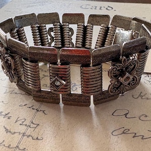 Könnte beinhalten: Silberfarbenes Armband mit rechteckigen und zylindrischen Gliedern. Das Armband hat dekorative florale Akzente und eine rautenförmige Verzierung. Der Schmuck im Vintage-Stil wird auf einem Vintage-Papierhintergrund präsentiert.