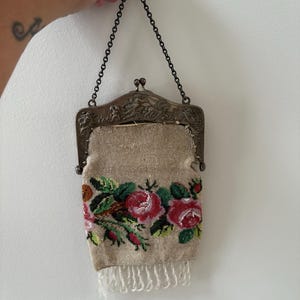 May include: An antique beaded purse with a metal frame and chain. The beige purse features a floral design with red and pink roses, green leaves, and white fringe at the bottom. The purse is held by a metal ring.