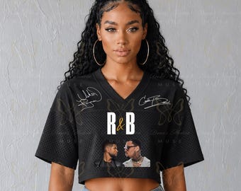 May include: A black cropped jersey with a V-neckline and short mesh sleeves. The front features the text R&B in white, two white signatures, and a graphic print of two men. The garment is paired with blue denim jeans, showcasing a casual aesthetic.