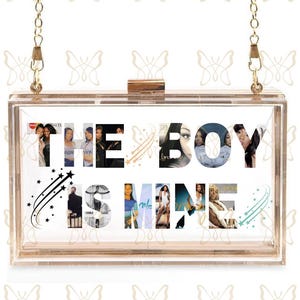 May include: A clear acrylic clutch with a gold chain strap. The front of the clutch features the words "THE BOY IS MINE" with images of women incorporated into the letters. Decorative star and trail accents are also present.