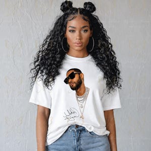 May include: A white short-sleeved t-shirt featuring a graphic print of a man with a beard wearing black sunglasses and layered silver chain necklaces. Below the graphic is a black cursive signature. The shirt is paired with blue denim jeans.