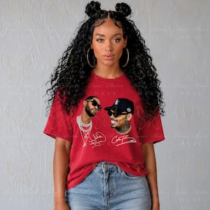 May include: A red short-sleeved t-shirt featuring a graphic print of two individuals wearing sunglasses and chains. The design includes white cursive signatures below each portrait. The shirt is paired with blue denim jeans.