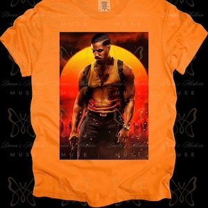 May include: Orange t-shirt with a graphic print of a man in a tank top and jeans, standing in front of a sunset. The man is holding a gun in his right hand and has a serious expression on his face.
